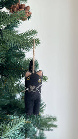 A playful black wool felt cat ornament hangs on a lush green Christmas tree. This handcrafted felt ornament features pointy peach ears, yellow eyes, and white whiskers, with a jute string attached for easy hanging.