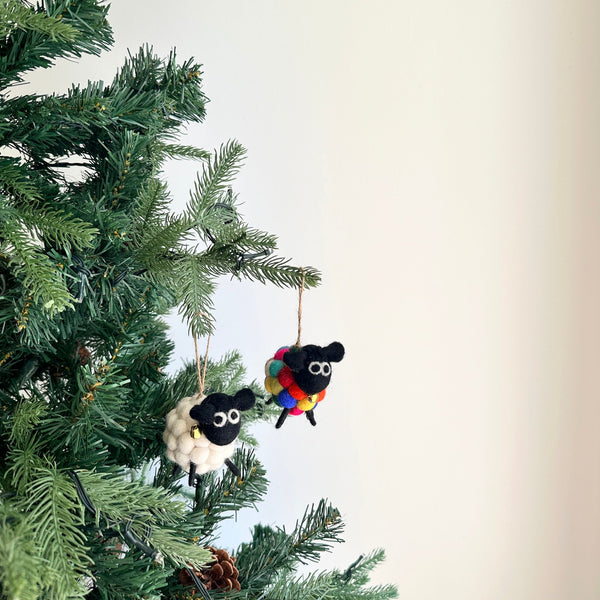 Two delightful wool felt pompom ball mini sheep Christmas ornaments hang from the vibrant green branches of an artificial Christmas tree. One mini sheep ornament has a white, pompom-like body with a small golden bell, while the other features a multi-colored felted body. Both sweet ornaments are detailed with black heads, thin black legs, and are suspended by a jute string.