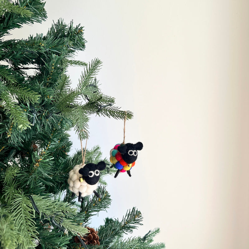 Two delightful wool felt pompom ball mini sheep Christmas ornaments hang from the vibrant green branches of an artificial Christmas tree. One mini sheep ornament has a white, pompom-like body with a small golden bell, while the other features a multi-colored felted body. Both sweet ornaments are detailed with black heads, thin black legs, and are suspended by a jute string.