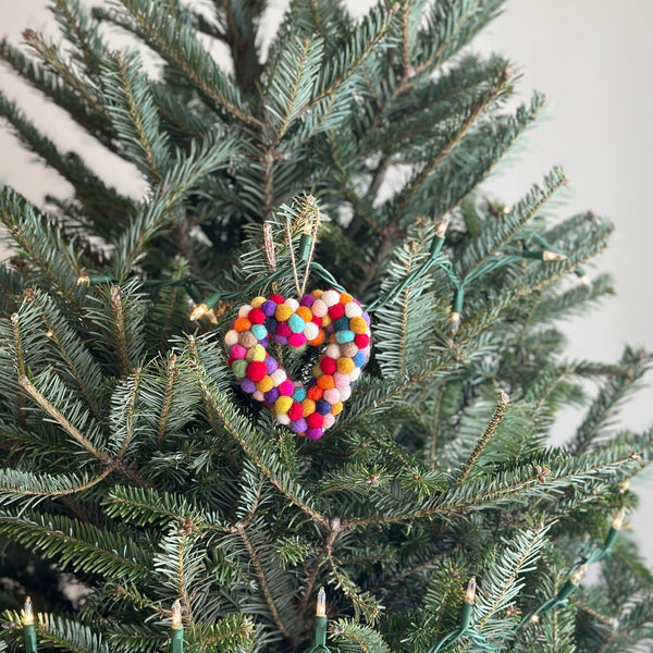 A Pompom Rainbow wool felt sheep ornament hangs on a lush green Christmas tree illuminated with warm white lights. This handmade felted sheep features a soft, textured body crafted from organic wool, adorned with small, colorful felt balls in red, blue, yellow, orange, purple, and green. A string for hanging is visible at the top, adding a cozy, joyful touch.