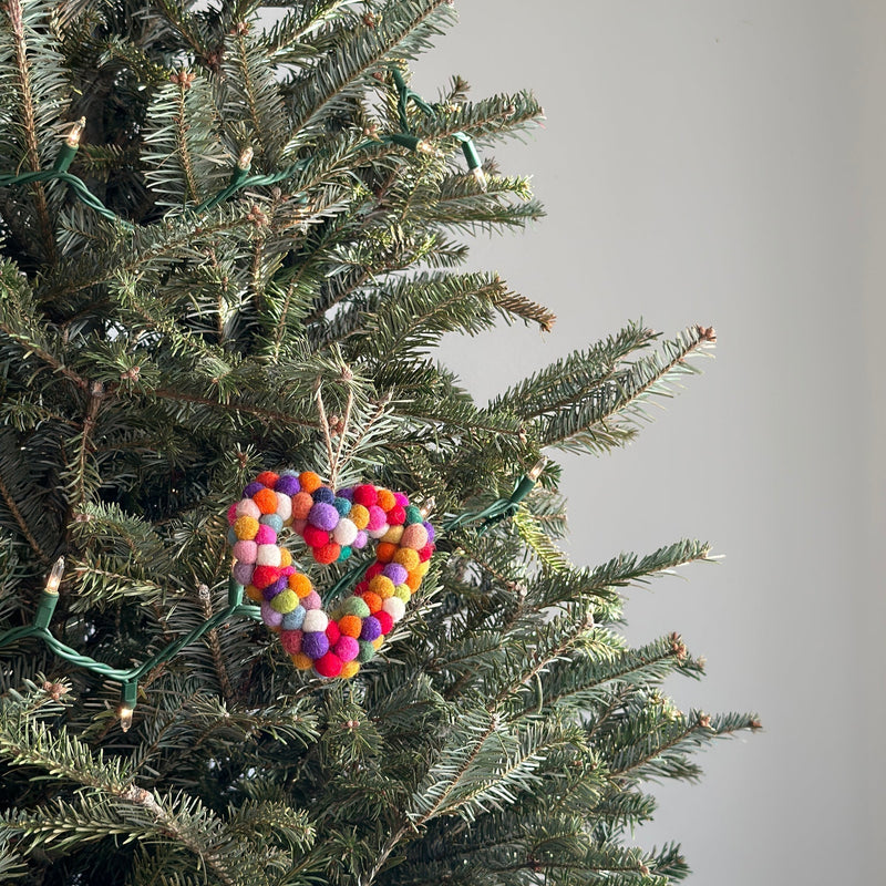 A wool felt pompom heart charm, crafted from colorful rainbow pompoms, hangs on a Christmas tree adorned with small electric lights. This unique Christmas ornament is suspended by a jute string, blending with the green needles of the festive tree.