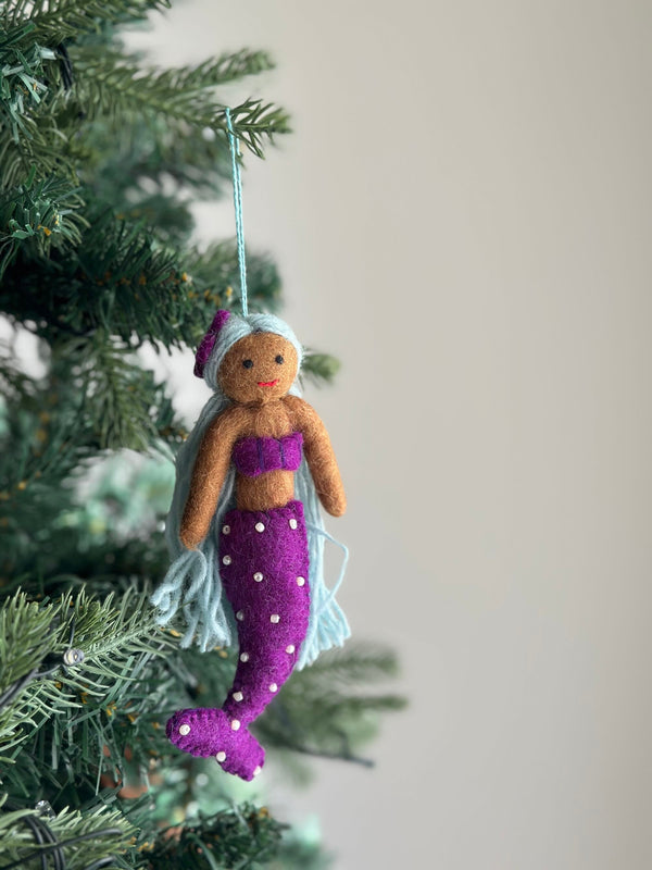 A handcrafted felt Pride Mermaid ornament hangs suspended by a light blue string on a lush, full Christmas tree against a soft light gray backdrop. This vibrant felt Pride Mermaid features a light brown upper body, a bold purple top, and a matching tail adorned with small white dots. Long, vibrant light blue hair flows from beneath its purple and light blue cap, creating a cheerful, whimsical, and joyful Christmas tree ornament.