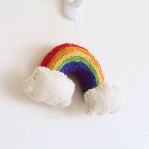 A vibrant handmade felt rainbow Christmas ornament hangs cheerfully from a white hook against a plain white wall. This 3D wool felt ornament features distinct bands of red, orange, yellow, green, blue, and purple. Fluffy white clouds anchor each end of the rainbow, adding to its three-dimensional and whimsical design. It is suspended by a thin, clear string.