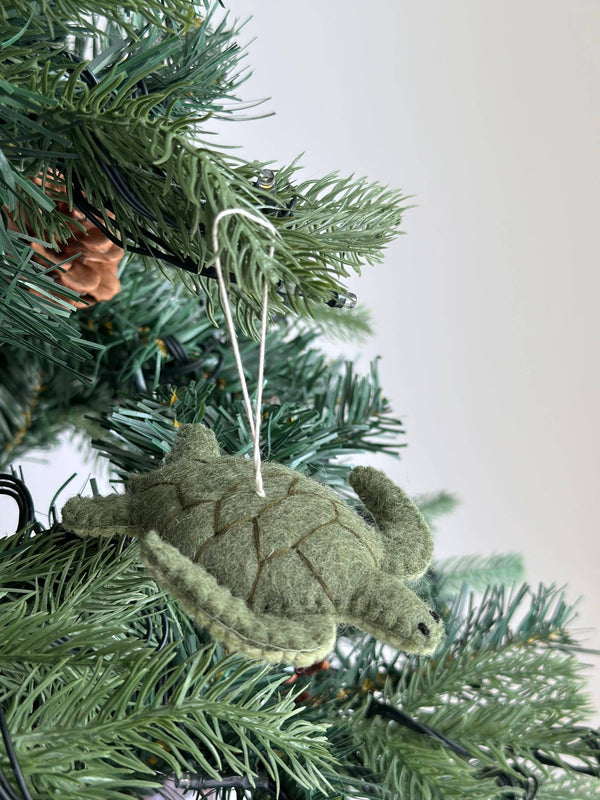 A textured light green wool felt sea turtle ornament hangs gracefully on a Christmas tree. Darker green stitching outlines its shell segments, and a white cotton string is attached for easy hanging, adding rustic charm to the holiday decor.