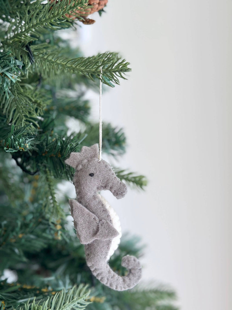 A gray and white wool felt seahorse ornament hangs gracefully on a lush green Christmas tree, set against a plain white wall, creating a heartwarming holiday atmosphere. This handcrafted felt ornament features a textured wool surface, a distinctive curled tail, and a small black eye, suspended by a thin cotton string.