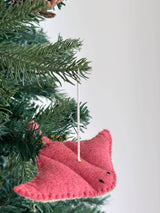 A pink wool felt stingray ornament hangs on a lush green Christmas tree, displayed against a bright white background. This charming sea animal felt ornament features a textured surface, meticulously stitched edges, small black stitched eyes, and a white cotton string for hanging.