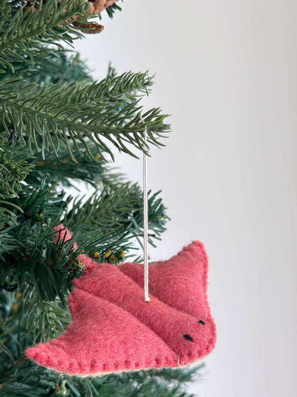 A pink wool felt stingray ornament hangs on a lush green Christmas tree, displayed against a bright white background. This charming sea animal felt ornament features a textured surface, meticulously stitched edges, small black stitched eyes, and a white cotton string for hanging.