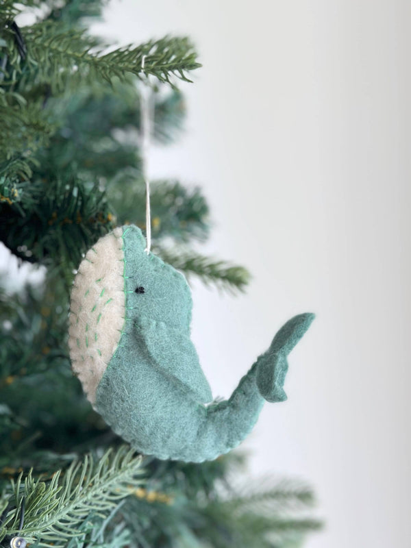A handcrafted wool felt whale ornament, part of a Sea Animals Theme, hangs on a pine Christmas tree. This light blue whale features a white belly with light green stitch details and is suspended by a white cotton string.