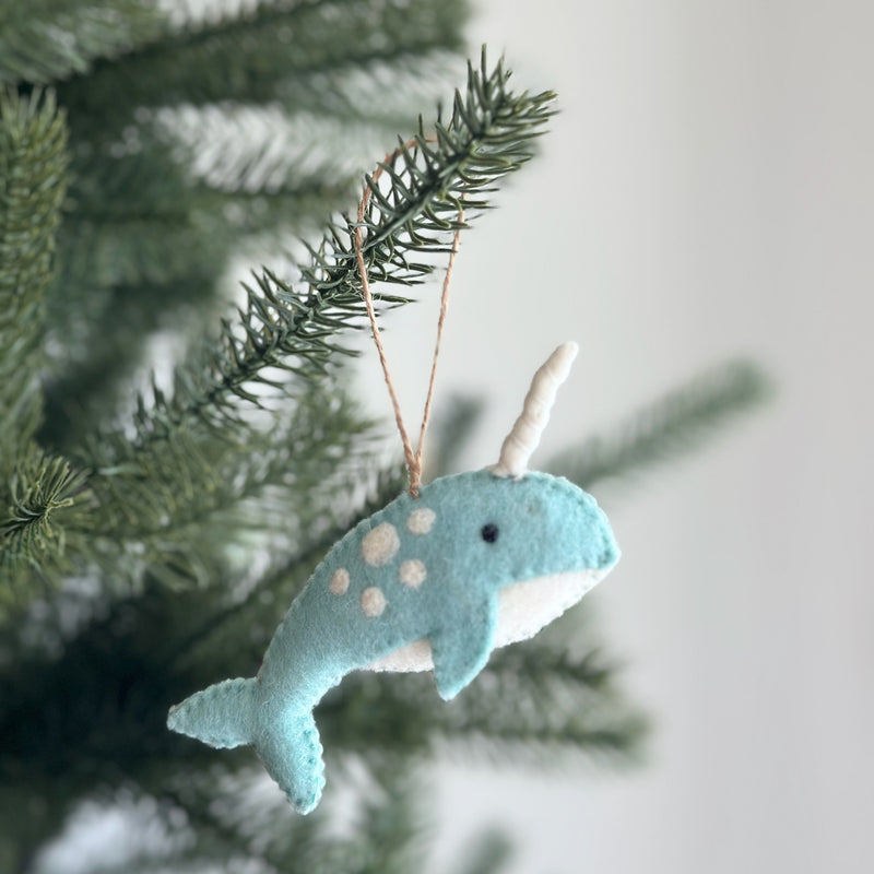 An ocean blue needle felted narwhal ornament, part of a unique Christmas ornament set, rests on a festive pine branch. This whimsical handmade felt narwhal ornament features white spots on its back, a soft white horn, and a delicate brown string for hanging, creating a cozy, cheerful holiday atmosphere.