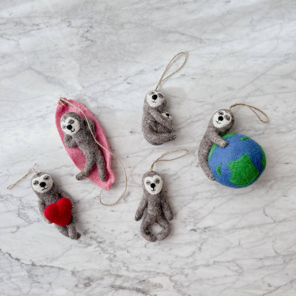 A charming wool felt sloth ornament, featuring a sloth on a pink felt hammock, is displayed on a marbled white surface. The handcrafted sloth has black eyes and a white face, appearing relaxed in its suspended jute string hammock. This unique ornament evokes a sense of whimsical leisure.