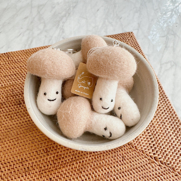 A Smiley Beige Mushroom felt ornament playfully rests in a light gray bowl, placed on a textured light brown woven rattan placemat. This handcrafted felt ornament features a soft beige, rounded cap and cylindrical body with a whimsical stitched smiley face. A silver string for hanging evokes a comfortable, nature-inspired feel.