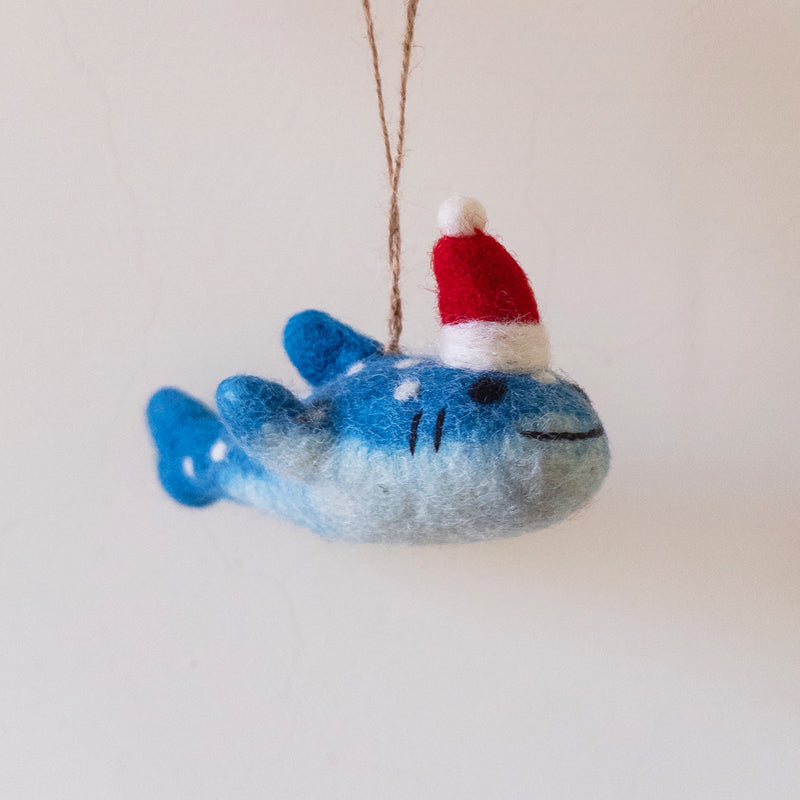 A Smiley Whale Shark felt ornament is suspended by a jute string against a plain, light background. This playful whale shark felt ornament features a gradient of blue hues, darker at the top and lighter towards the bottom, adorned with white spots. It has a simple stitched smile and black gills, creating a cute and approachable appearance.