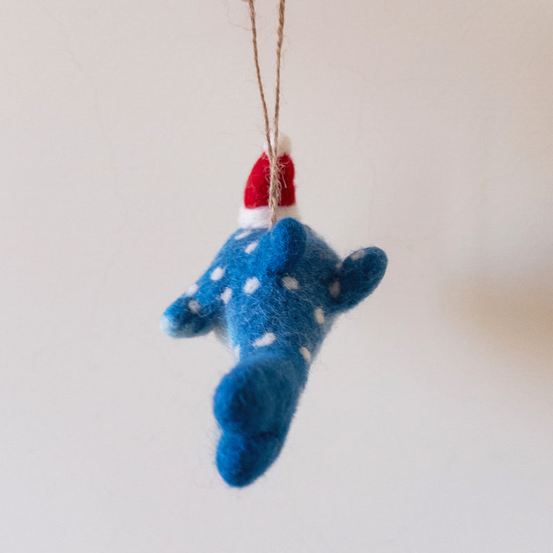 A unique felt whale shark ornament hangs against a light background. This whimsical blue whale shark ornament features a friendly smile and white polka dots, topped with a festive red and white Santa hat. A jute string is attached for easy display.