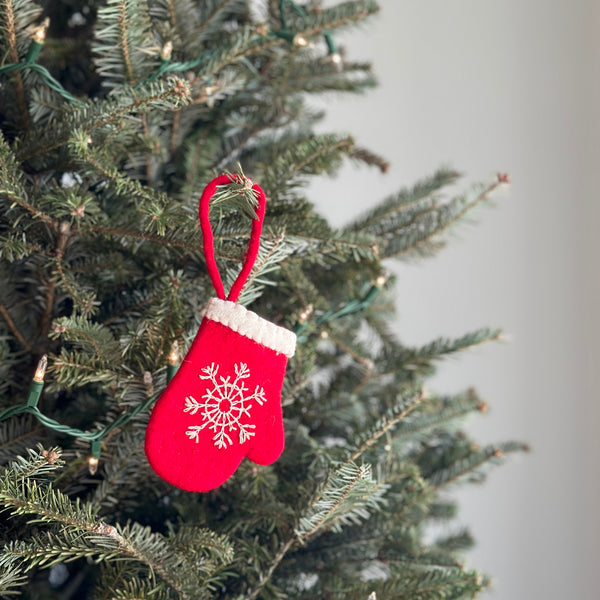 A felt snowflake glove ornament hangs from a Christmas tree adorned with delicate lights, perfectly displayed for the holiday season. This handcrafted wool felt ornament is bright red with a fluffy white trim and a detailed white hand-embroidered snowflake on the front. A red cotton string is attached for easy hanging, adding to its cozy and festive charm.