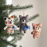 Multiple Stitched Cat Holding Fish wool felt ornaments hang from a Christmas tree branch, adding cozy charm. Each hand-stitched cat ornament features unique color patterns, such as a white cat with black and orange stripes and a gray cat with blue-gray stripes, both holding a blue fish. Intricate embroidered details adorn these playful handcrafted felt ornaments, each suspended by a jute string.