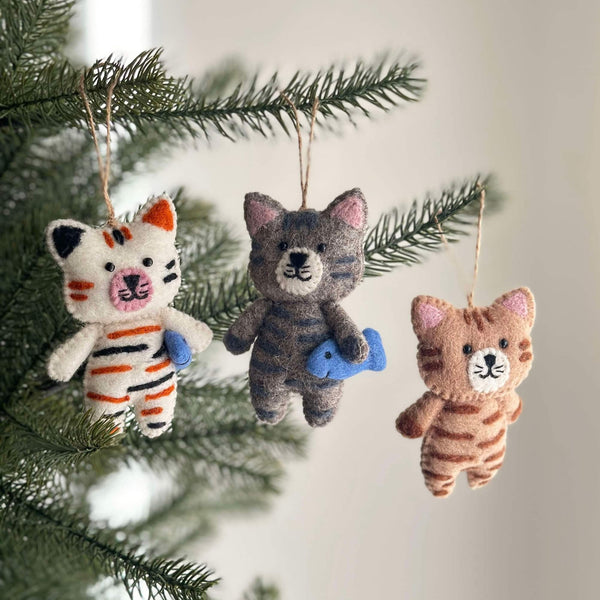 Multiple Stitched Cat Holding Fish wool felt ornaments hang from a Christmas tree branch, adding cozy charm. Each hand-stitched cat ornament features unique color patterns, such as a white cat with black and orange stripes and a gray cat with blue-gray stripes, both holding a blue fish. Intricate embroidered details adorn these playful handcrafted felt ornaments, each suspended by a jute string.