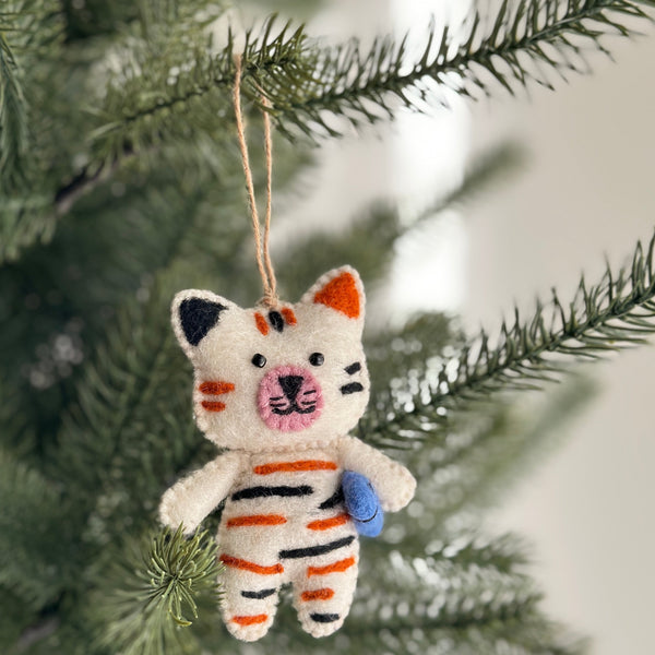A playful wool felt cat ornament holding a fish hangs on a lush green Christmas tree. This unique Christmas ornament features a soft white body with black and orange stripes. Hand-stitched details define the cat's face, while a blue fish is clutched in its paws. A jute string allows for easy hanging.