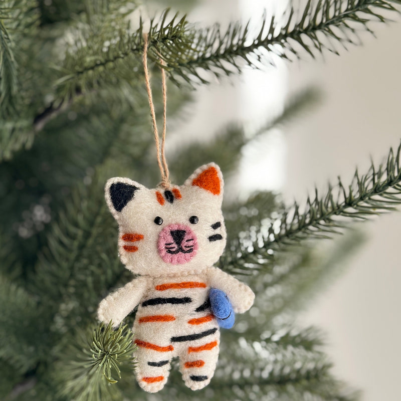 A playful wool felt cat ornament holding a fish hangs on a lush green Christmas tree. This unique Christmas ornament features a soft white body with black and orange stripes. Hand-stitched details define the cat's face, while a blue fish is clutched in its paws. A jute string allows for easy hanging.