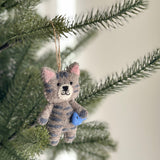 The charming wool felt stitched cat ornament, holding a blue fish, hangs on a green Christmas tree. This handmade grey cat features beige wool accents, pink inner ears, and dark grey stripes. It has black eyes, a stitched mouth and whiskers, and is suspended by a natural jute string, adding a festive and cozy touch.