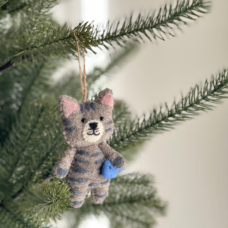 The charming wool felt stitched cat ornament, holding a blue fish, hangs on a green Christmas tree. This handmade grey cat features beige wool accents, pink inner ears, and dark grey stripes. It has black eyes, a stitched mouth and whiskers, and is suspended by a natural jute string, adding a festive and cozy touch.
