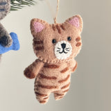 A charming stitched wool felt cat ornament hangs from a pine branch. This light brown wool felt cat ornament features dark brown stripes, small pointed ears with pink inner details, a white muzzle with black whiskers, and tiny black eyes. A jute string is attached for easy display.