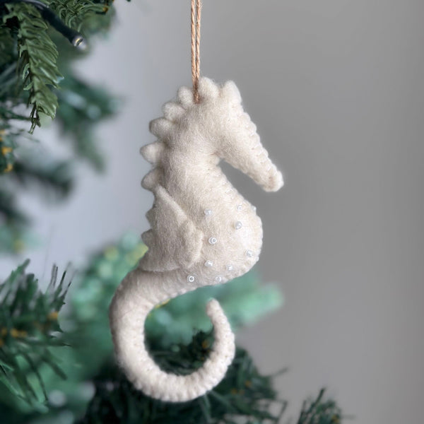A white seahorse felt ornament hangs on a subtly lit Christmas tree branch. This handcrafted felt ornament features a textured white felt surface embellished with small, clear beads. A jute string is attached, completing its decorative charm for a classic Christmas setting.