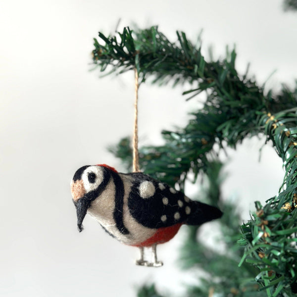 A felt woodpecker ornament hangs on an artificial Christmas tree branch against a bright backdrop. This detailed handcrafted felt ornament has prominent black and white markings on its wings and head, accented with red at its rear and gray legs. It creates a joyful and handmade aesthetic.