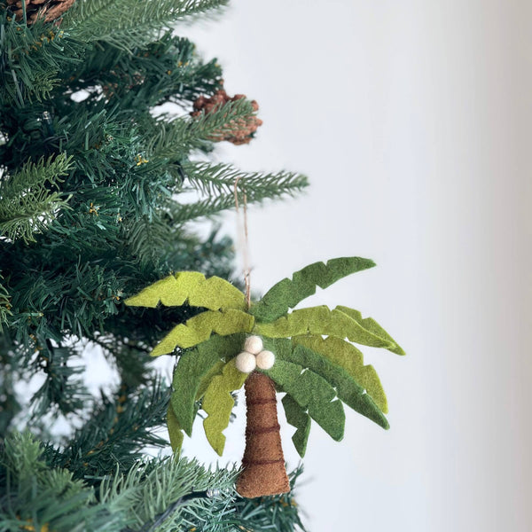 Bringing tropical warmth, a felt palm tree with coconut ornament is nestled among the evergreen branches of a Christmas tree. This unique felt ornament features vibrant green leaves, a sturdy brown trunk, and three white felted coconuts clustered beneath the fronds, complete with a jute string for hanging.