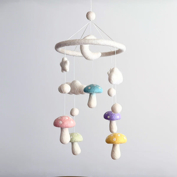 A Felt Pastel Mushroom Baby Mobile hangs against a pale gray backdrop, creating a whimsical and dreamy scene. This charming wool felt baby mobile features pastel mushrooms in pink, light green, blue, purple, and yellow, each with white polka dots. Fluffy white wool felt stars, clouds, and a gentle crescent moon dangle at varying lengths from a soft white felt ring, suspended by thin white strings.