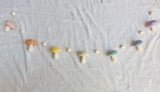 A 4-foot felt garland featuring soft, handcrafted wool felt mushrooms in pastel colors, including blush, honey, mint, lilac, and ivory, is laid out on a white surface. The whimsical garland consists of multiple toadstools and balls, evoking feelings of enchantment and playfulness. Made from 100% wool felt, this unique garland is perfect for nurseries, playrooms, or woodland-themed parties.