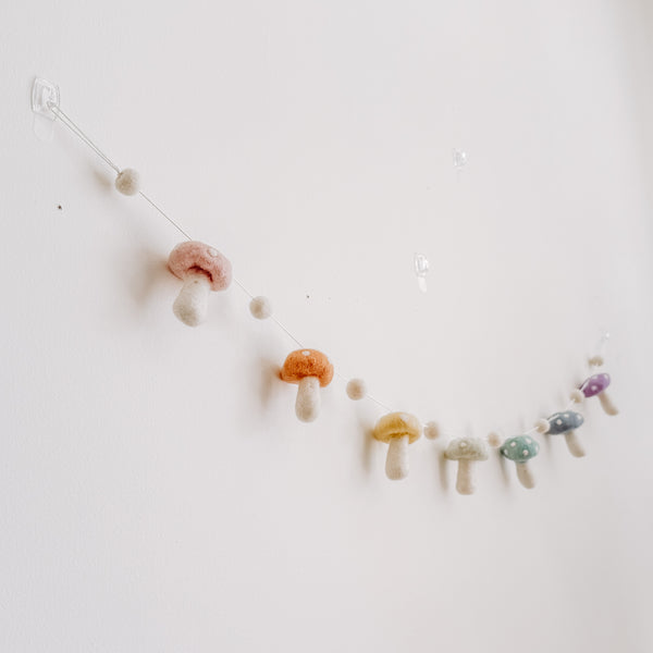 A felt pastel mushroom garland is displayed on a light, neutral wall, evoking a whimsical and cozy atmosphere. The handcrafted felt garland is attached with transparent hooks and features alternating soft pastel wool felt balls and wool felt mushrooms. Each wool felt mushroom, in blush, honey, mint, and lilac hues, has white dots on top.