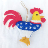 A handcrafted wool felt patriotic chicken ornament is displayed against a white backdrop. This unique Christmas ornament features a blue body with white stars, a red and white striped tail, and sunny yellow details on its head and feet. A jute string is attached for hanging.
