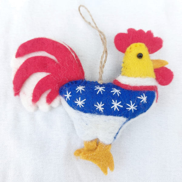 A handcrafted wool felt patriotic chicken ornament is displayed against a white backdrop. This unique Christmas ornament features a blue body with white stars, a red and white striped tail, and sunny yellow details on its head and feet. A jute string is attached for hanging.
