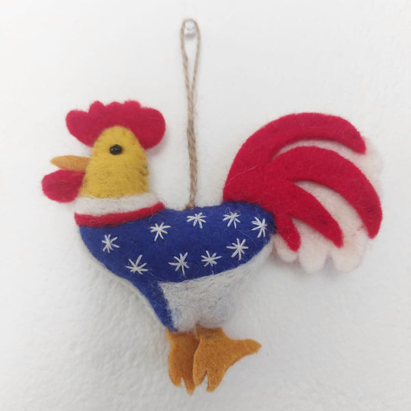 A handmade wool felt patriotic rooster ornament, hanging from a jute string, is displayed against a plain white background. This festive rooster features a bold red crest and tail feathers, a blue body adorned with white stars, and sunny yellow accents on its head and feet.
