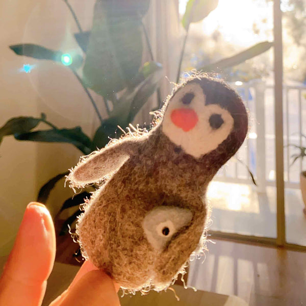 A handmade felt penguin finger puppet resting on a human hand, set against a cozy indoor background featuring a plant and natural light streaming through a window. The penguin is brown with a cheerful expression, carrying a small fish in its beak. This wool felted finger puppet is 3D designed and perfect for storytelling.