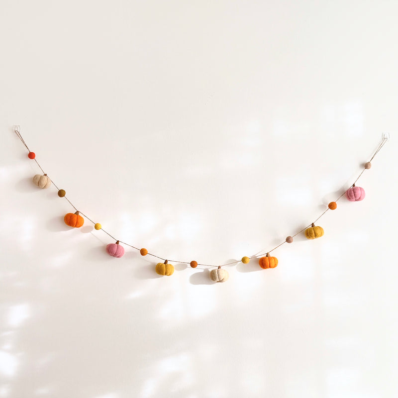 A handcrafted wool felt pumpkin garland is displayed, gently curved, against a clean white wall. This 48-inch autumn-themed garland features eight felted pumpkins in various fall colors, including orange, tan, light pink, and golden yellow. The pumpkins are strung together with jute thread, creating a cozy and charming seasonal decoration.