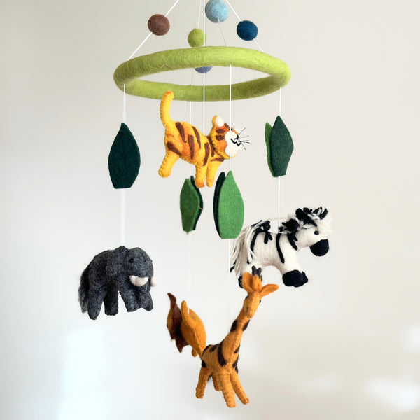 A felt safari animal baby mobile hangs from a ceiling in a nursery setting, featuring hand-felted animals like lions, elephants, zebras, giraffes, and tigers with distinct patterns and colors. The mobile is perfect for a child's room or nursery.