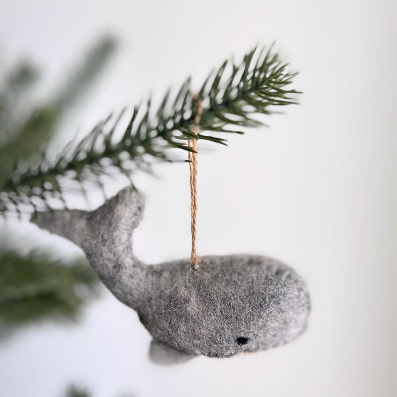 A gray whale felt ornament, part of the Felt Sea Animal Ornaments collection, hangs gracefully from a pine branch against a soft white background. This wool felt gray whale features a streamlined body with an upturned tail, a single black eye, and textured gray felt, ready for display.