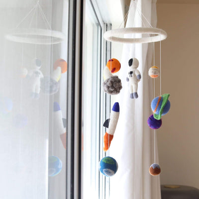 A colorful wool felt spaceman baby mobile hangs suspended in front of a bright window with white curtains, creating a soft, dreamy atmosphere. The mobile features a white felt ring from which a white spaceman, colorful planets, an orange and blue rocket, and a gray asteroid are suspended. Each needle-felted figure, crafted from 100% New Zealand wool, is detailed and textured, offering a whimsical, three-dimensional look.