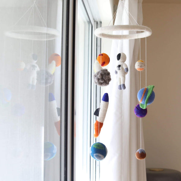 A handcrafted wool felt spaceman baby mobile hangs from a ceiling hook, suspended above a white curtain backdrop. The mobile features colorful space-themed elements, including planets, stars, and a spaceship made of 100% New Zealand wool felt. The design exudes a playful, whimsical mood, perfect for a child's room or nursery.