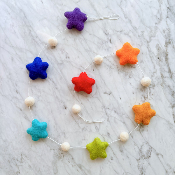 A cheerful wool felt star garland rests on a marble surface. This vibrant garland features seven soft, tactile felt stars in a rainbow of purple, blue, teal, green, red, orange, and yellow. White felt balls are evenly spaced between each star, all connected by a thin white string.