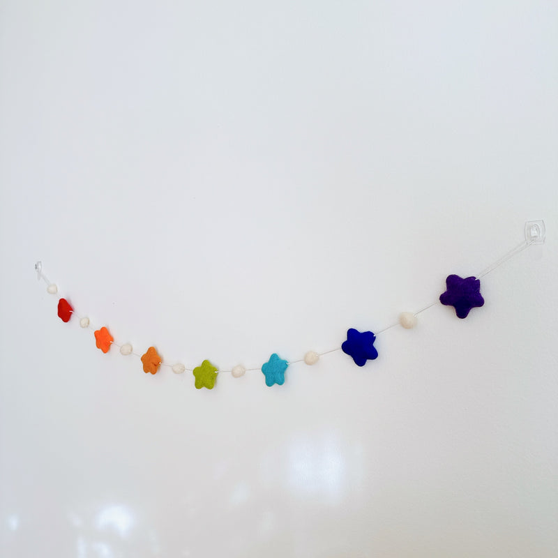 A vibrant felt star garland is displayed on a plain white wall, creating a cheerful atmosphere. This playful felt garland features seven rainbow-colored felt stars, including red, orange, yellow, green, light blue, dark blue, and purple. The stars are evenly spaced along a thin white string, interspersed with small white felt balls.