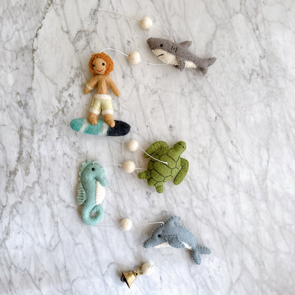 A playful Felt Surfer Garland rests on a marbled white surface. This handmade felt garland features a tan felt surfer on a blue and white striped surfboard, a gray felt shark with a white underbelly, a green felt turtle, a teal felt seahorse, and a gray felt dolphin. Each charming ocean-themed element is connected by a string with small white felt balls, finished with a copper bell.
