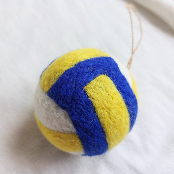A vibrant, round handmade felt volleyball ornament rests on a soft, white fabric background. Crafted from wool felt, this ornament features classic yellow, blue, and white panels inspired by official volleyballs. A natural jute string loop is visible at the top, ready for hanging.