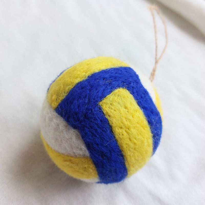 A vibrant, round handmade felt volleyball ornament rests on a soft, white fabric background. Crafted from wool felt, this ornament features classic yellow, blue, and white panels inspired by official volleyballs. A natural jute string loop is visible at the top, ready for hanging.