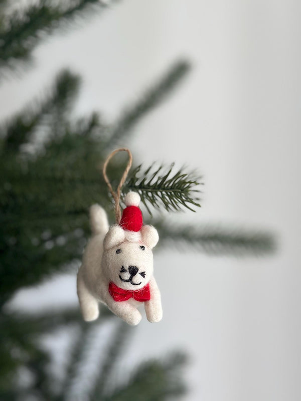 A charming handmade felt white cat Christmas ornament, wearing a festive red Christmas hat and a matching red bow tie, hangs from a pine branch by a jute string. The delightful felt cat ornament is presented against a blurred background of a Christmas tree and a plain white wall, ready for holiday display.
