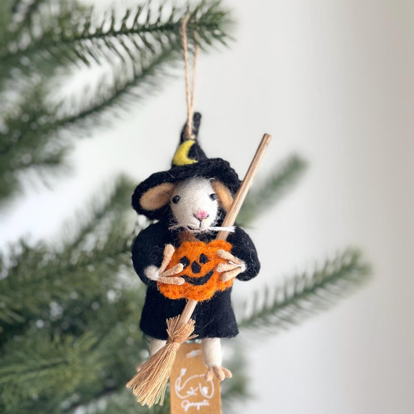 A playful, handmade wool felt wizard mouse ornament, holding a miniature orange pumpkin and wooden broom, hangs on a lush green Christmas tree. The white mouse wears a pointy black hat with a yellow crescent and a black cloak. It features tiny white whiskers, beige ears, and pink paws, with a jute string for hanging.