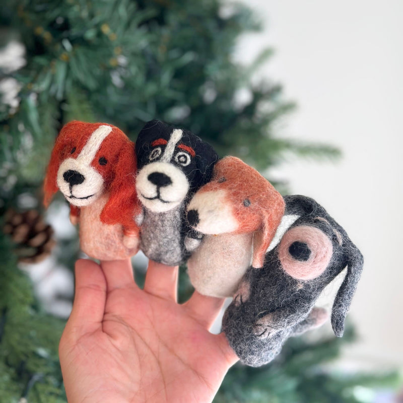 Felt dog finger puppets resting on a festive Christmas tree background, surrounded by ornaments and lights. The puppets feature various breeds with distinct fur textures, colors, ears, tails, and facial expressions. Each felt puppet is handmade in Nepal, showcasing unique designs that evoke feelings of warmth, comfort, and creativity.