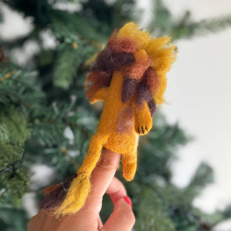 A fluffy wool felt lion finger puppet, handcrafted ethically in Nepal, is set against a blurred green Christmas tree, creating a festive scene. This unique felt toy features a bright yellow body, a fluffy mane with gradients of brown, tan, and gold, and black felt claws on its front paws.