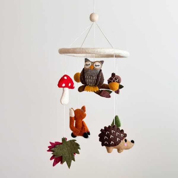 A whimsical hanging mobile featuring felt animals and objects, suspended from a circular felt hoop by strings. The mobile showcases an owl perched on a branch, surrounded by a fox, hedgehog, mushroom, acorn, and pine tree, all crafted from handcrafted wool felt. This delightful mobile brings the enchanting forest world to life in a nursery setting.